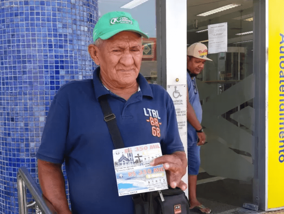 Foto mostra home idoso, de camisa azul, boné verde, com bilhetes de loteria na mão para vender.