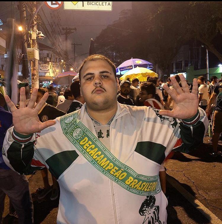 Giovanni tomoatti torcedor palmeirense na rua fazendo o sinal de 10 coma mão, no peito uma faixa escrito "decacampeão brasileiro"