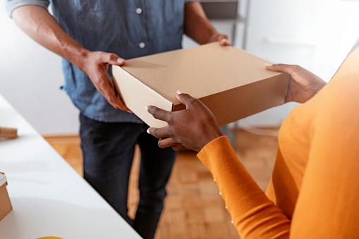 Um homem de traje azul entrega de um caixote para uma mulher que está vestindo uma blusa laranja