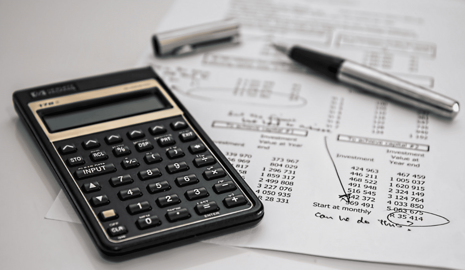 Foto mostra uma calculara, caneta e folhas em cima de uma mesa.