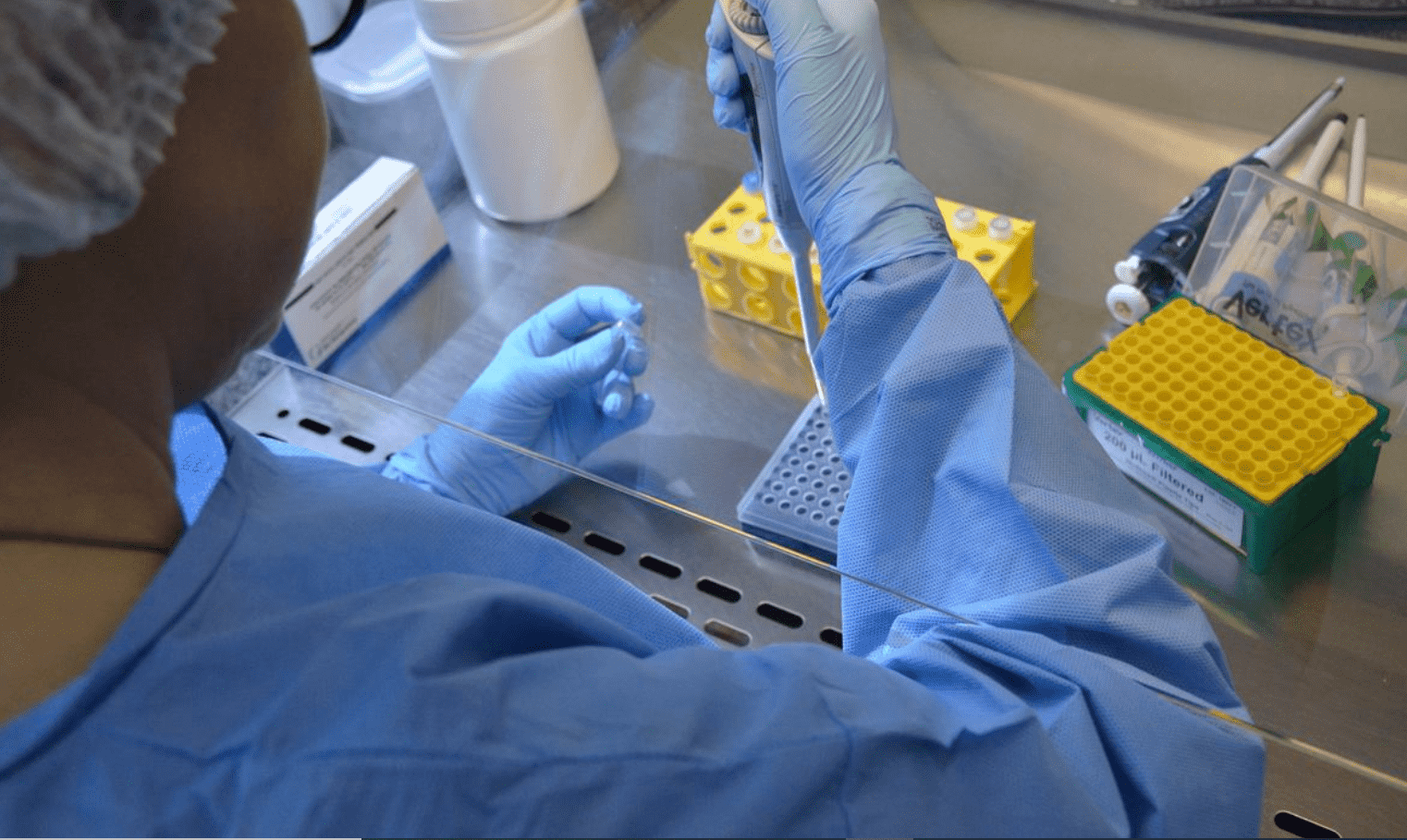 Foto mostra pessoa com roupa de proteção no laboratório de costas mexendo em fracos.