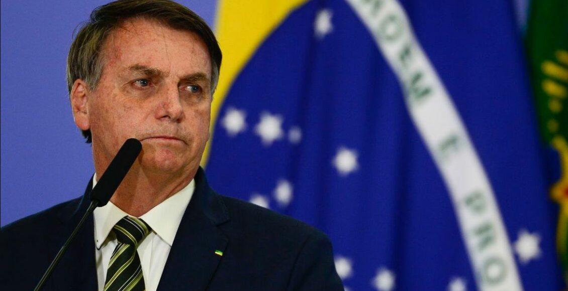 Foto mostra presidente Jair Bolsonaro alinhado a esquerda do enquadramento, vestido de terno, diante de um microfone e com olhar direcionado a direita (sua esquerda). Bandeira do Brasil se encontra ao fundo.