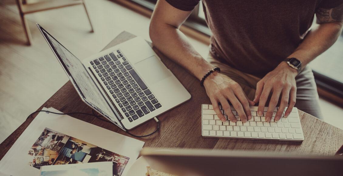 Foto aproximada mostra homem digitando sobre teclado de computador sobre mesa de madeira, com notebook ao lado.